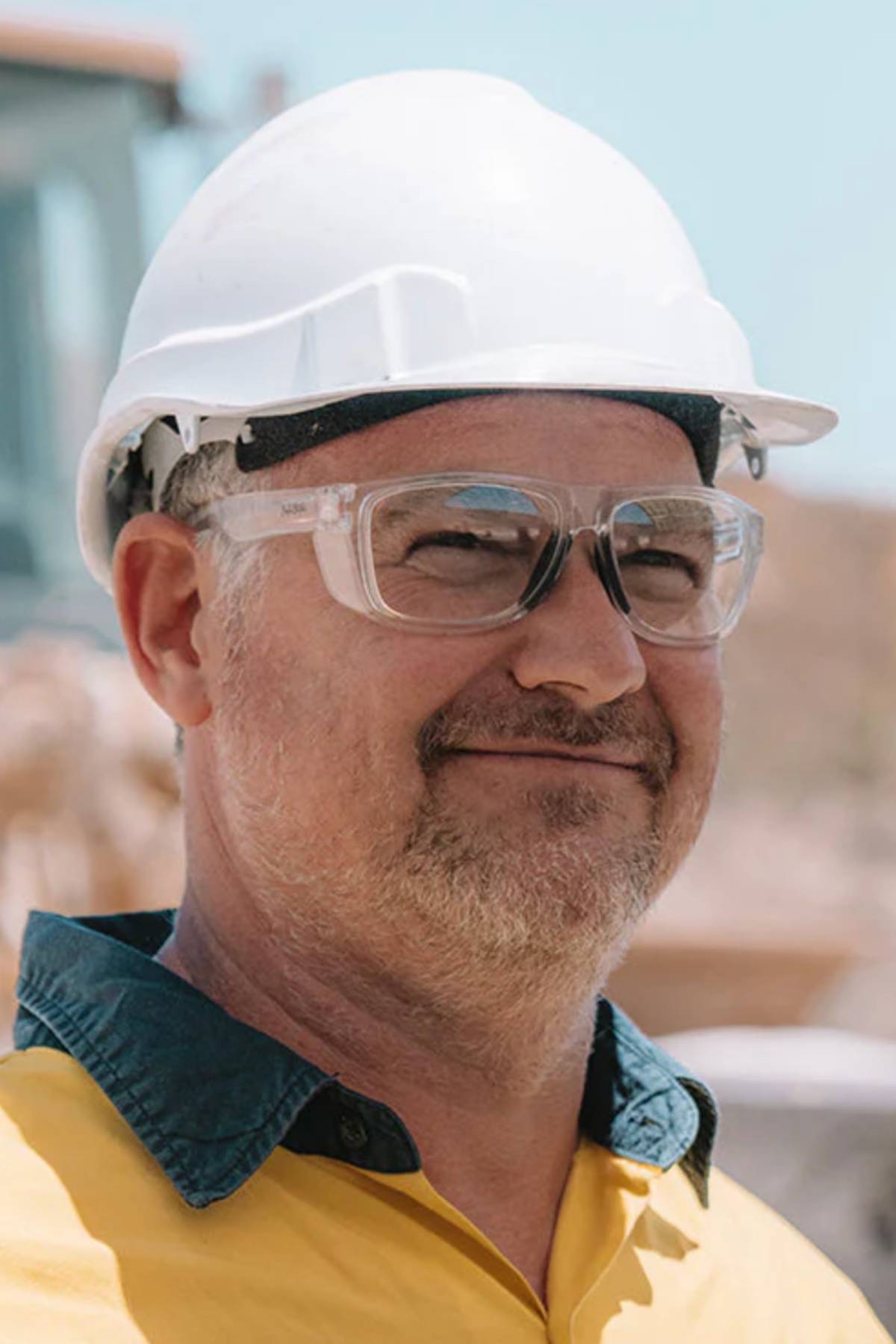 A middle-aged man in a yellow shirt with a dark collar stands outdoors at a construction site, wearing SafeStyle Rapids Clear Frame Clear Lens safety glasses.