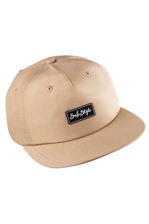The Safestyle Worksite to Weekend Essentials Cap is a beige snapback with a flat brim, made from recycled polyester. It features a black "SafeStyle" patch, ventilation eyelets, and offers UPF 50+ sun protection.