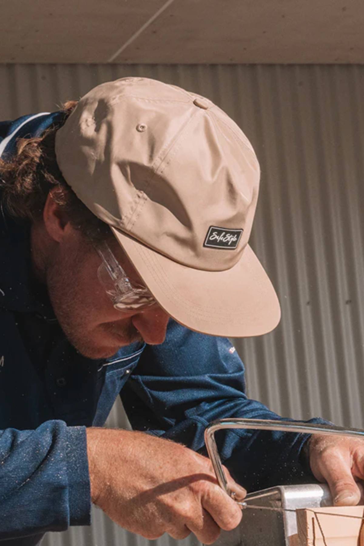 Wearing the Safestyle Worksite to Weekend Essentials Cap and safety glasses, a person uses a hand saw to cut wood, with a corrugated metal wall behind them.