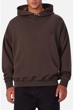 A person wearing a loose-fitting, dark brown Industrie The Sandblast Hoodie crafted from 100% cotton, featuring a front pocket, paired with black pants and standing against a plain white background for an effortlessly laid-back style.