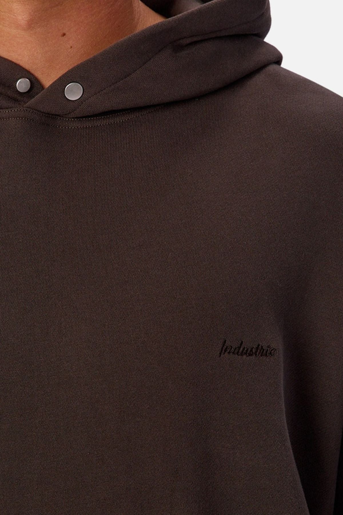 A close-up shows a person wearing the Industrie The Sandblast Hoodie, made from 100% cotton in dark brown with silver buttons and "Industrie" embroidered in black on the chest. Only their lower face, neck, and upper torso appear in the image, emphasizing the hoodie's relaxed style.