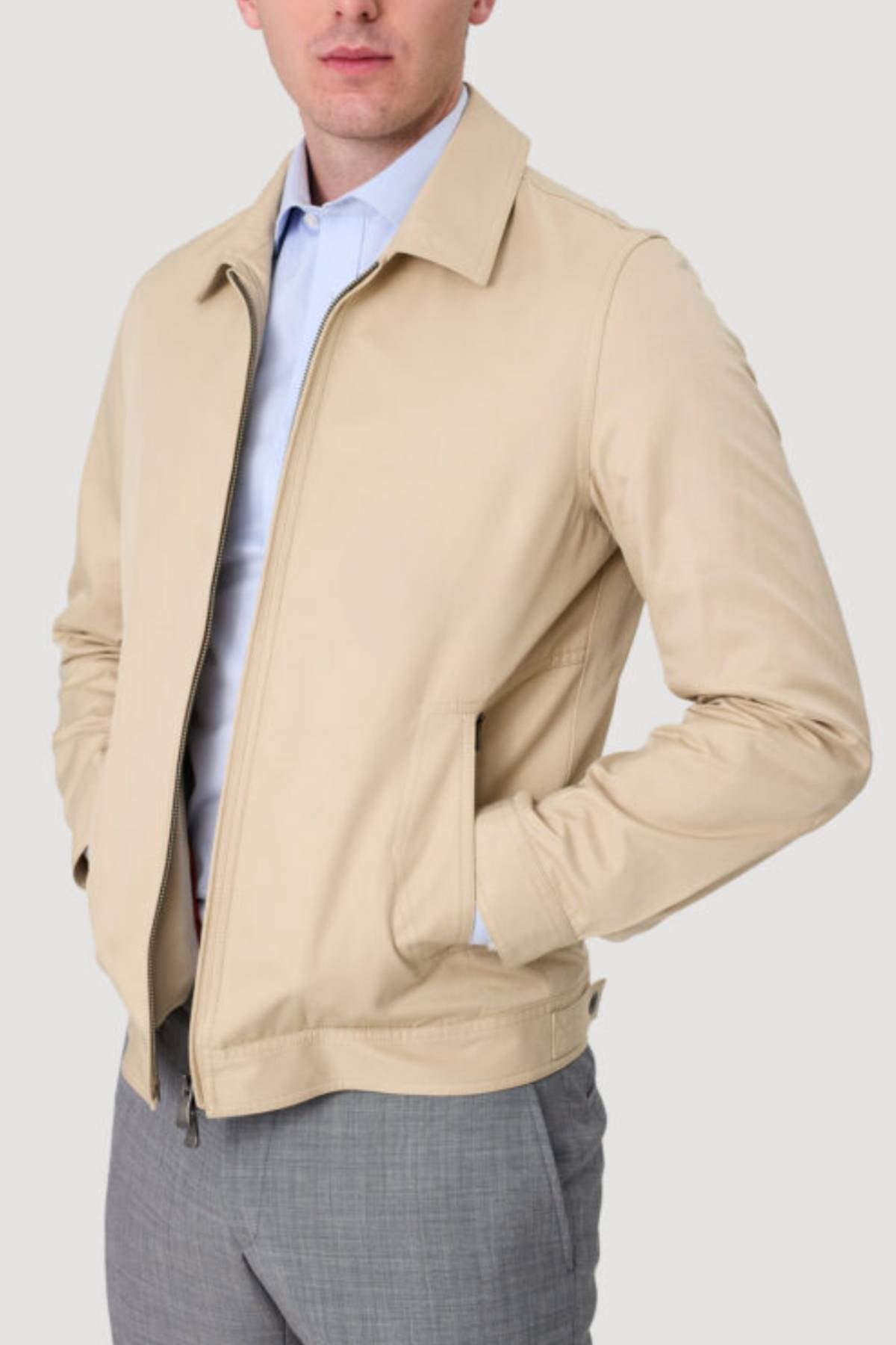 A man stands with his hands in the pockets of the Savile Row James Zip Front Jacket, crafted from 100% cotton, layered over a light blue dress shirt and gray trousers against a plain light background.