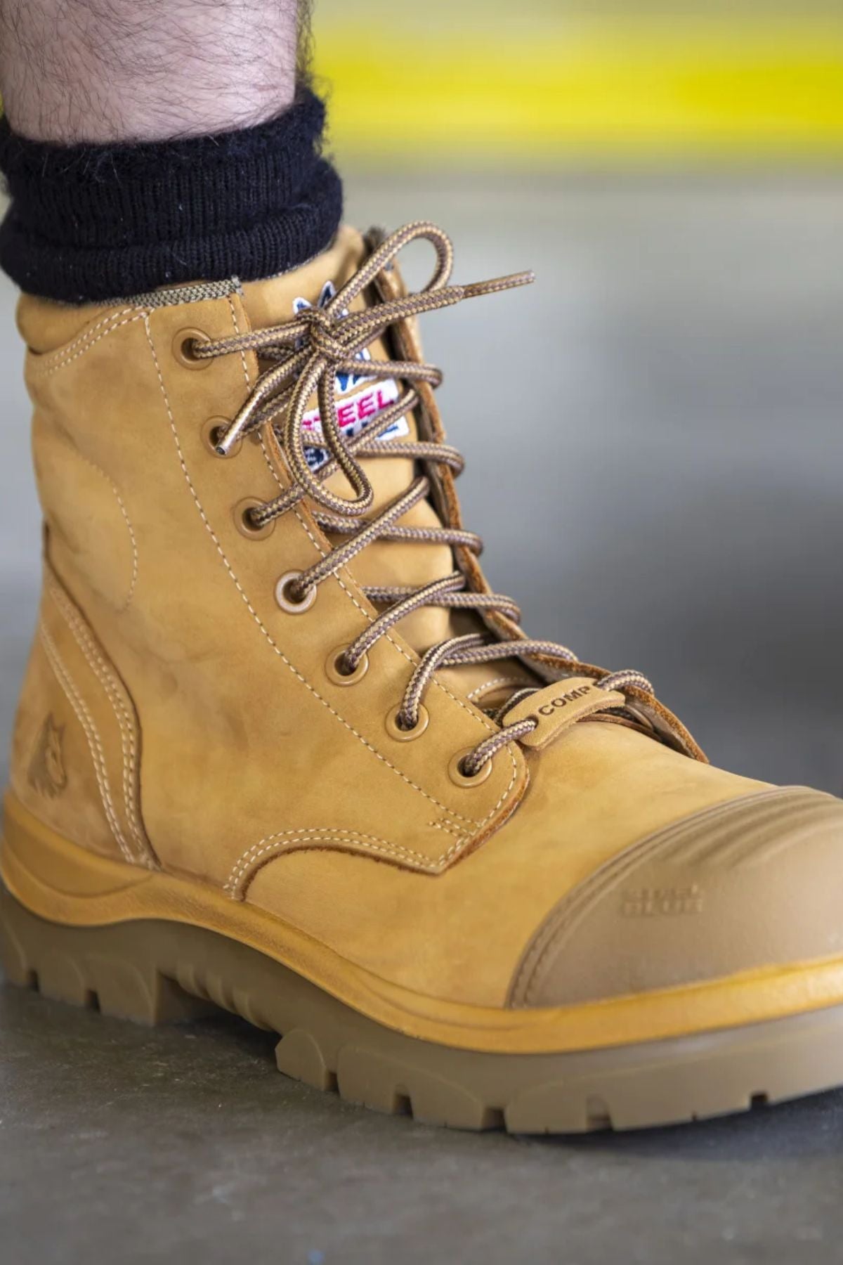A close-up of a person’s foot wearing Steel Blue Argyle Zip Scuff Cap Boot Composite Toe in tan, featuring a rugged sole, black sock, and visible brand labels, standing on a smooth gray surface.