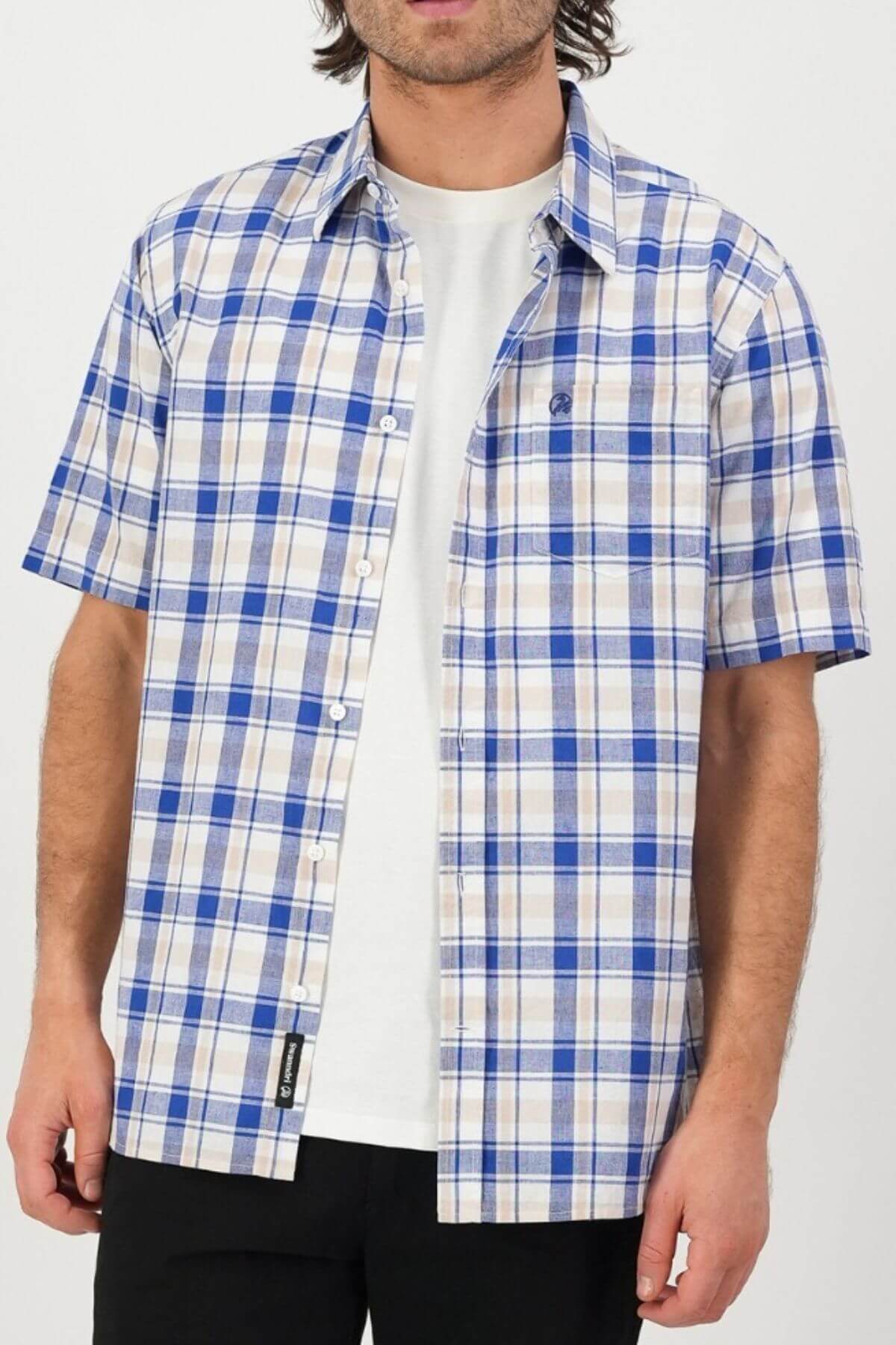 A person wears a Swanndri Coleridge Short Sleeve Shirt in blue and beige plaid over a white T-shirt, paired with black pants for a casual yet stylish look. The person's face isn't visible in the image.