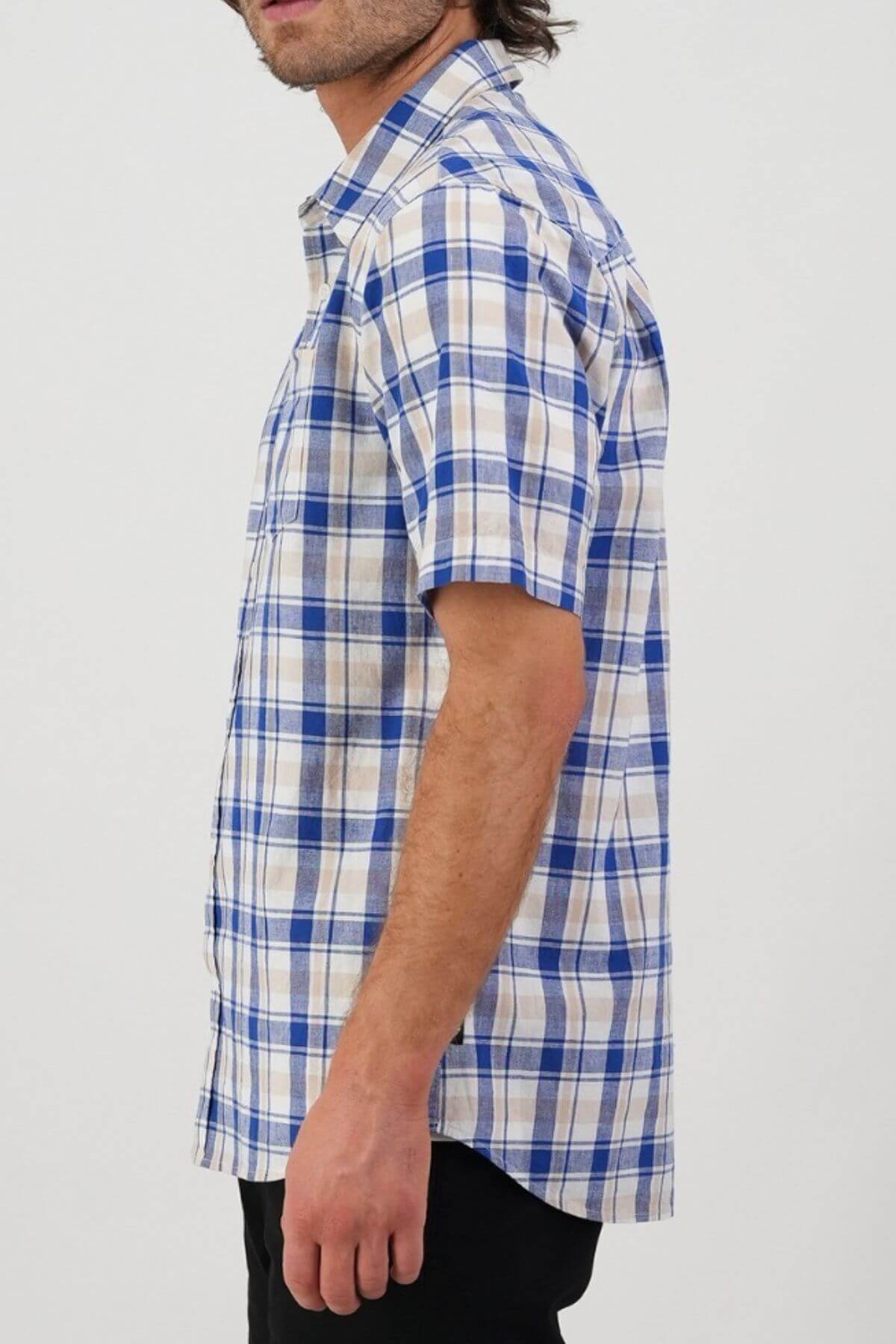 Wearing the Swanndri Coleridge Short Sleeve Shirt in a blue and white plaid, a person stands sideways against a plain light background. The shirt is buttoned up and paired with black pants, focusing attention on their upper attire.