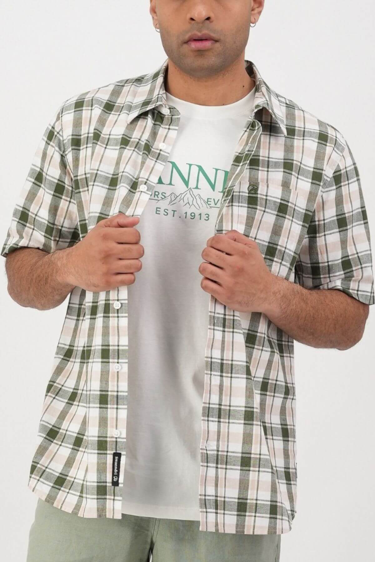 A man wears a white graphic tee layered with the Swanndri Coleridge Short Sleeve Shirt in a green and beige plaid cotton-linen blend, holding the sides for a relaxed vibe—perfect casual style on a plain white background.