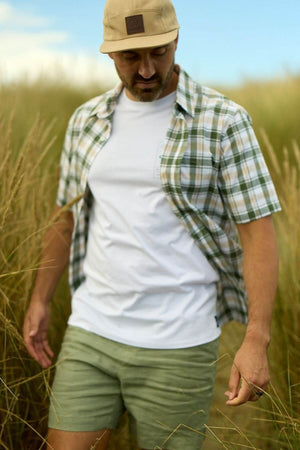 A man wearing a beige cap, Swanndri Coleridge Short Sleeve Shirt, and green shorts strolls through tall grass in a field on a sunny day—ideal for any casual outing.