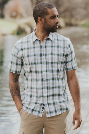 A man with short hair and a beard stands outdoors by the water, wearing a Swanndri Coleridge Short Sleeve Shirt (green and white plaid cotton-linen blend) and tan cargo shorts, looking left with one hand in his pocket—ideal for casual outings.