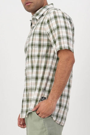 A man stands sideways with one hand in his pocket, wearing a Swanndri Coleridge Short Sleeve Shirt in green, beige, and white plaid with light green pants against a plain white background—ideal for any casual occasion.