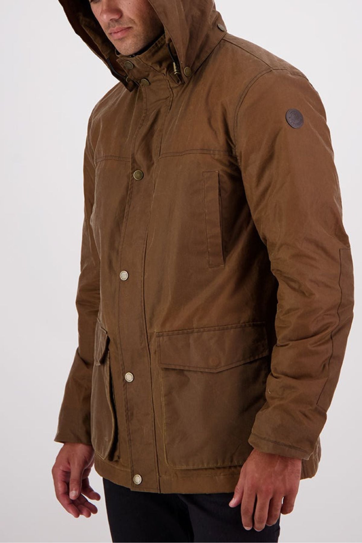 Man wearing the Swanndri Evans Flat v2 Oilskin Jacket, a brown waxed cotton hooded coat with front pockets and button closures. The fabric is subtly water-resistant. He stands against a plain light background, face partially visible as he looks downward.