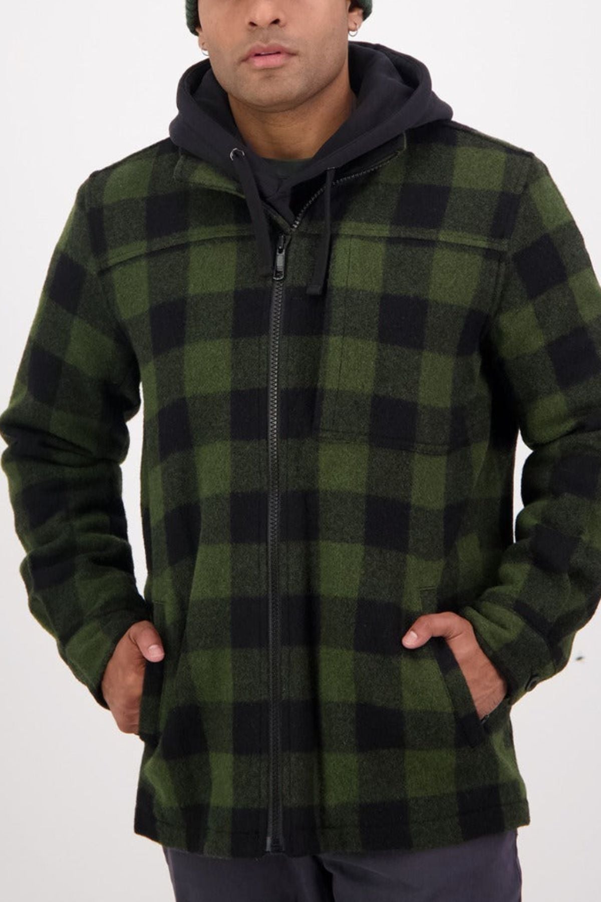 A person wears a green and black plaid Swanndri Hudson Jacket, made from New Zealand wool, over a black hoodie with hands in pockets, standing against a plain white background—ideal for stylish outdoor wear.