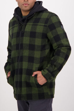 A person wears the Swanndri Hudson Jacket in green and black plaid, made from New Zealand wool, layered over a black hoodie with dark pants, hands in pockets—ideal for stylish outdoor wear against a light background.