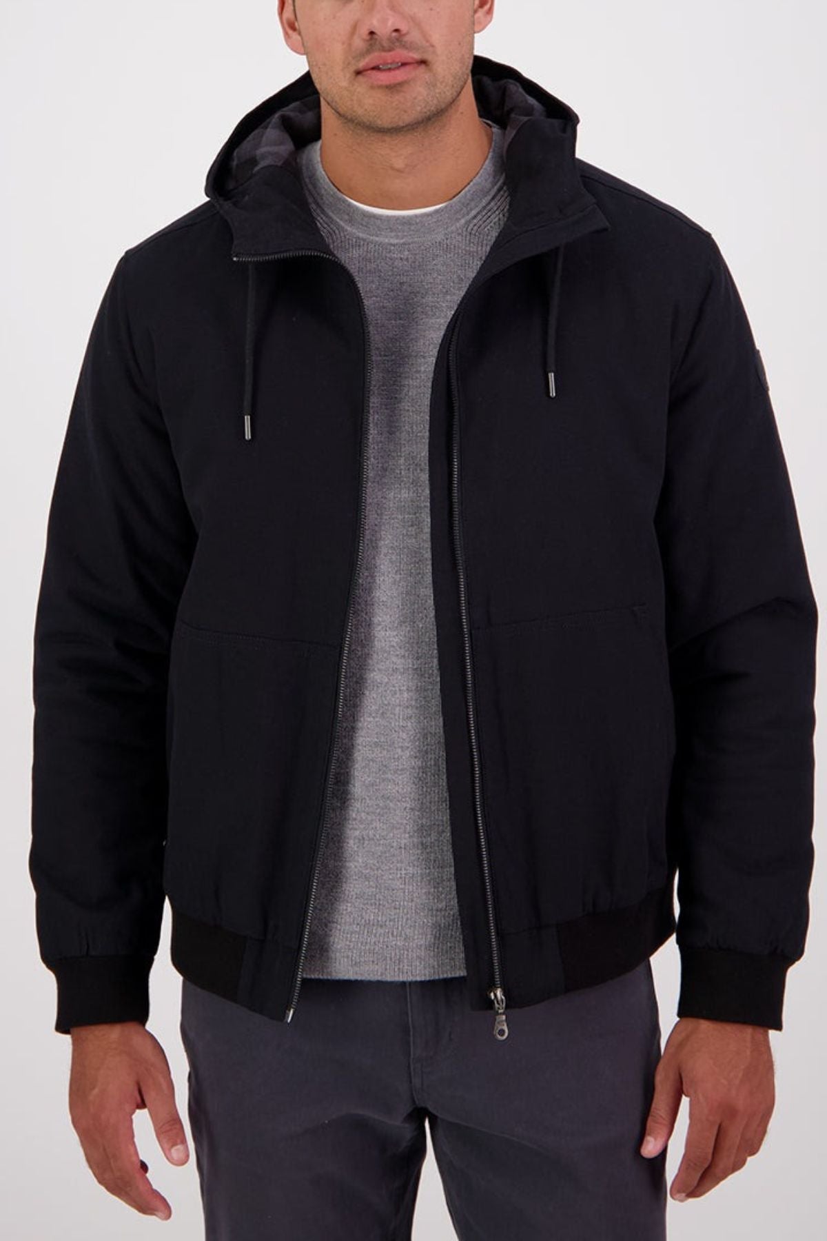 A man stands facing forward with his hands at his sides, wearing a Swanndri Whitlow v2 Canvas Hoodie over a gray sweater and dark gray pants against a plain light background.