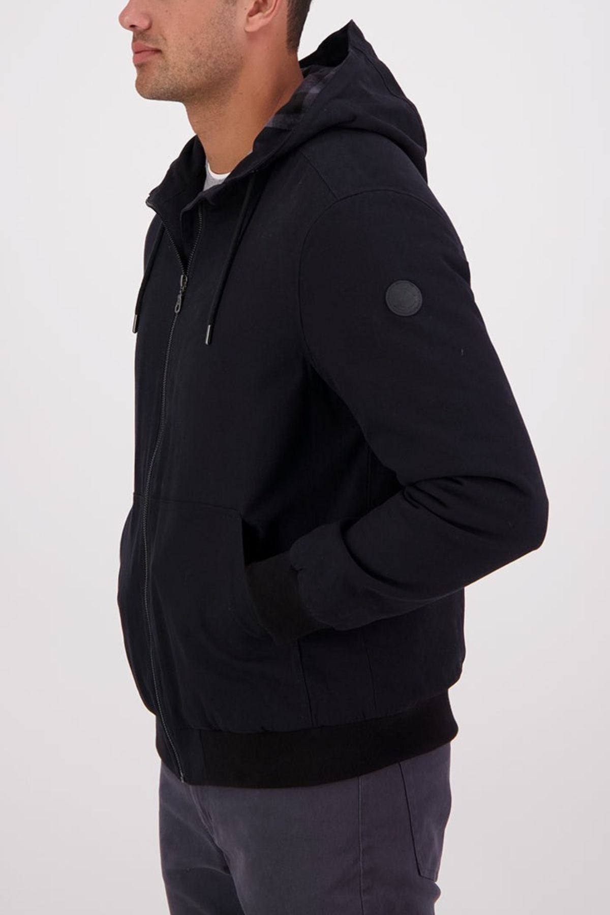 A man stands sideways against a plain light background, hands in the pockets of a black Swanndri Whitlow v2 Canvas Hoodie.