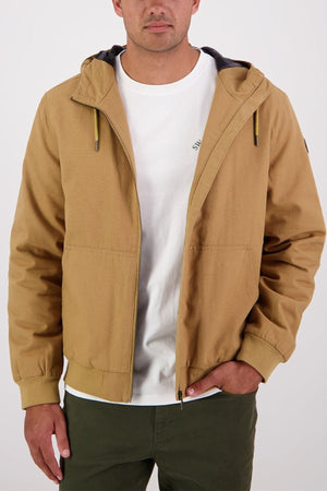 A man wears a Swanndri Whitlow v2 Canvas Hoodie—tan cotton canvas—over a white t-shirt and dark green pants, standing with one hand in his pocket against a plain background.