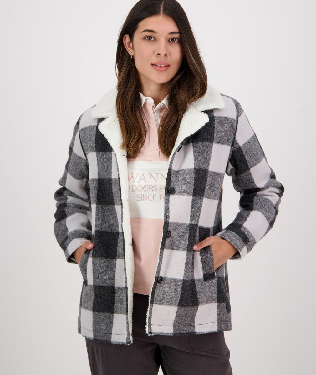 A woman is standing against a plain background wearing the Swanndri Capitola Sherpa Jacket, which features a black, white, and grey checkered pattern and a soft, white fuzzy lining. She has her hands in the jacket pockets. She has long, dark hair and is looking directly at the camera with a neutral expression.