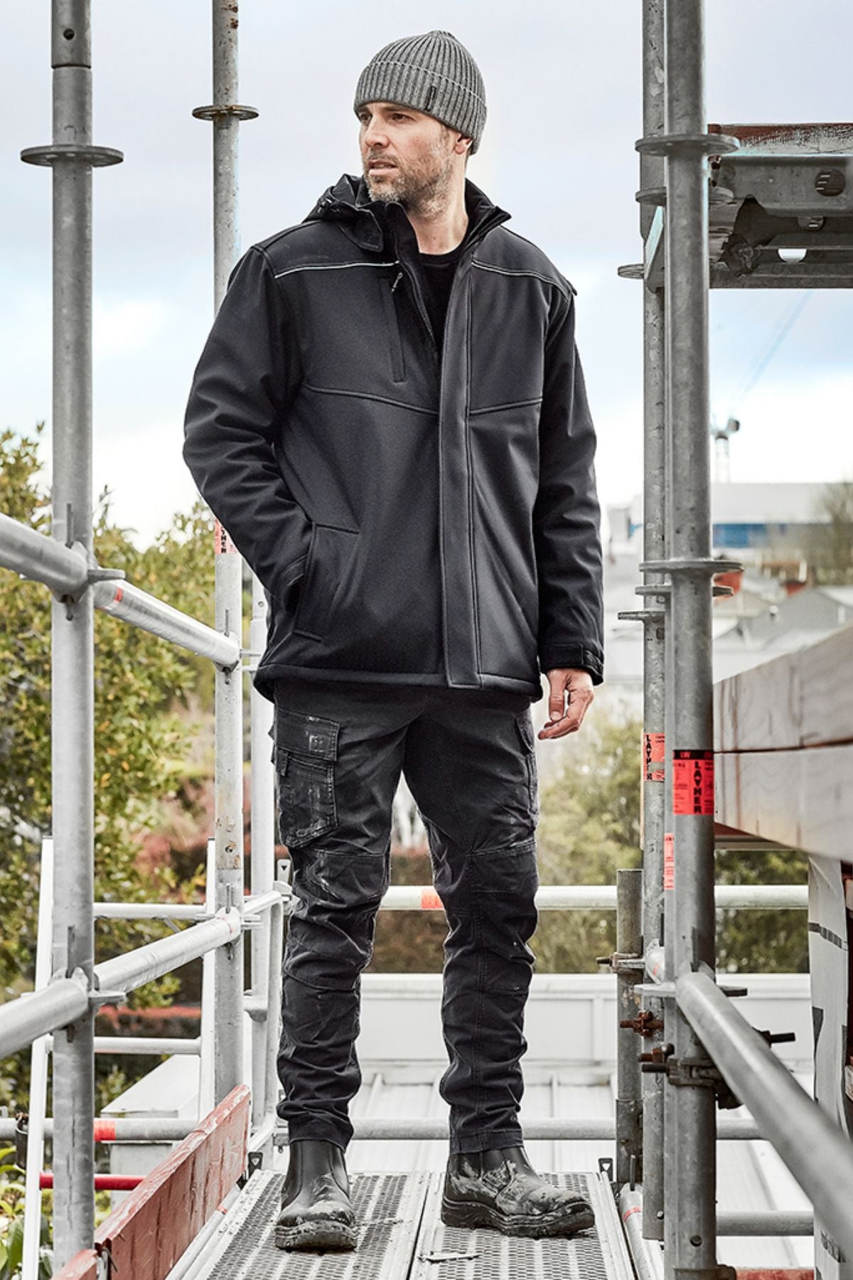 A man in a gray knit beanie, Syzmik Antarctic Softshell Jacket (black), black cargo pants, and black boots stands on an outdoor metal scaffold, looking to the side with trees and buildings in the background.