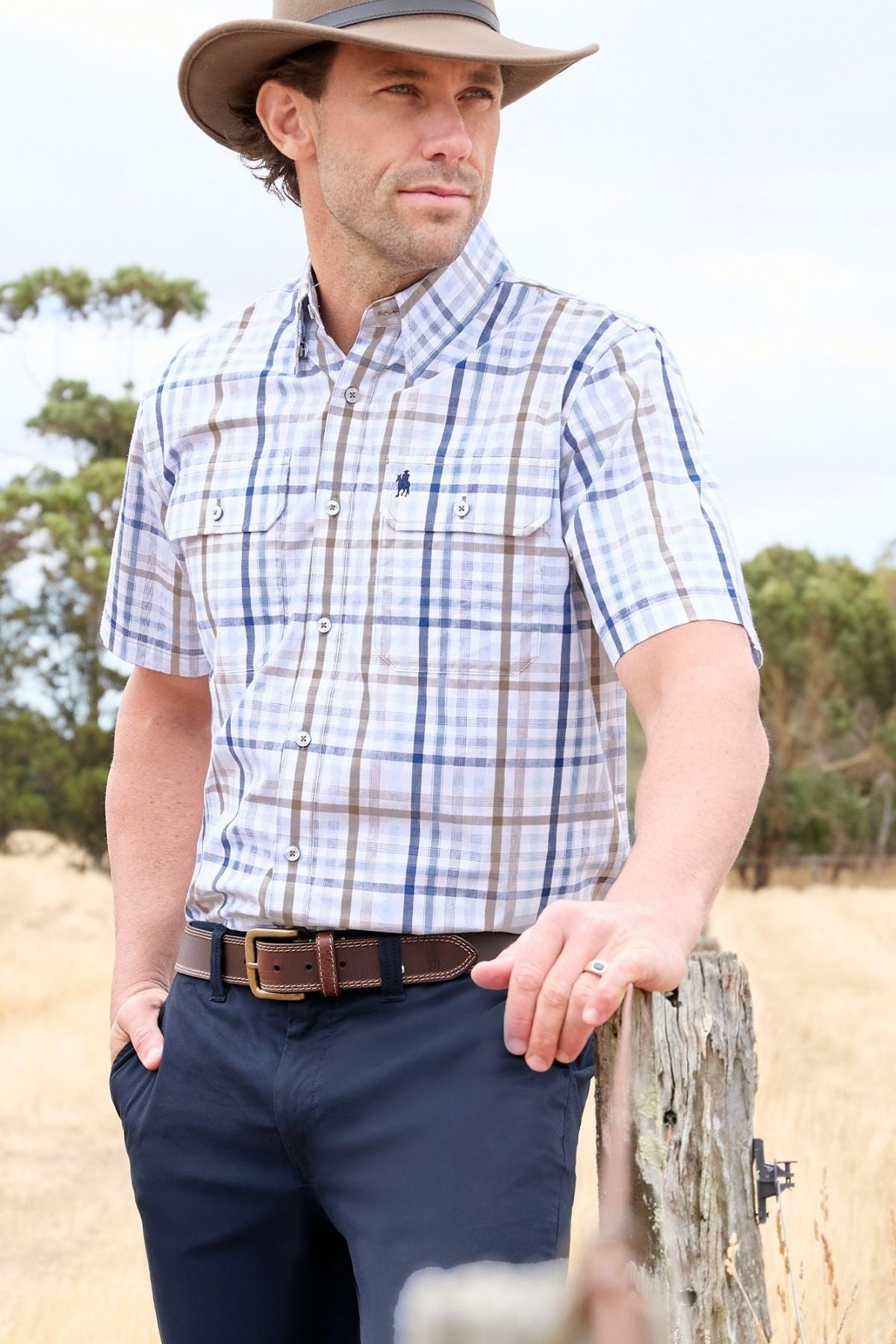 A man wearing the Thomas Cook Alfie Short Sleeve Shirt, navy pants, and a wide-brim hat leans on a wooden fence in a dry, grassy outdoor setting with trees in the background.