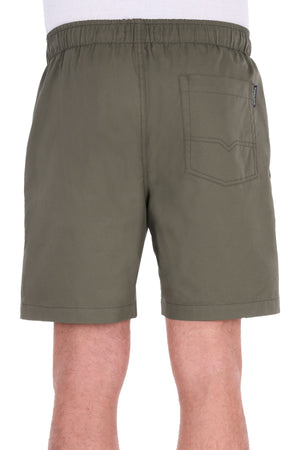A person is shown from the back wearing the Thomas Cook Darcy Short in olive green. The shorts, made from 100% cotton, have an elasticated waist and a single patch pocket on the right side. Visible in the image are the person’s legs and a small part of a white shirt.
