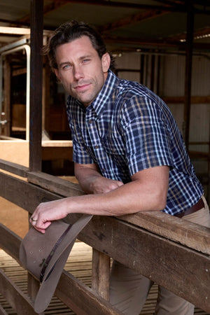 A man in a Thomas Cook Finley Short Sleeve Shirt leans on a wooden fence inside a barn, holding a cowboy hat in one hand and gazing at the camera with a relaxed expression. Crafted from 100% cotton, this classic workwear shirt ensures all-day comfort.