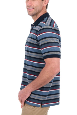 A man stands against a white background, turned slightly to the side and smiling, dressed in khaki pants and a Thomas Cook Glenn Polo—a short-sleeve polo shirt made from soft cotton pique with bold horizontal stripes in dark, light, and red tones.