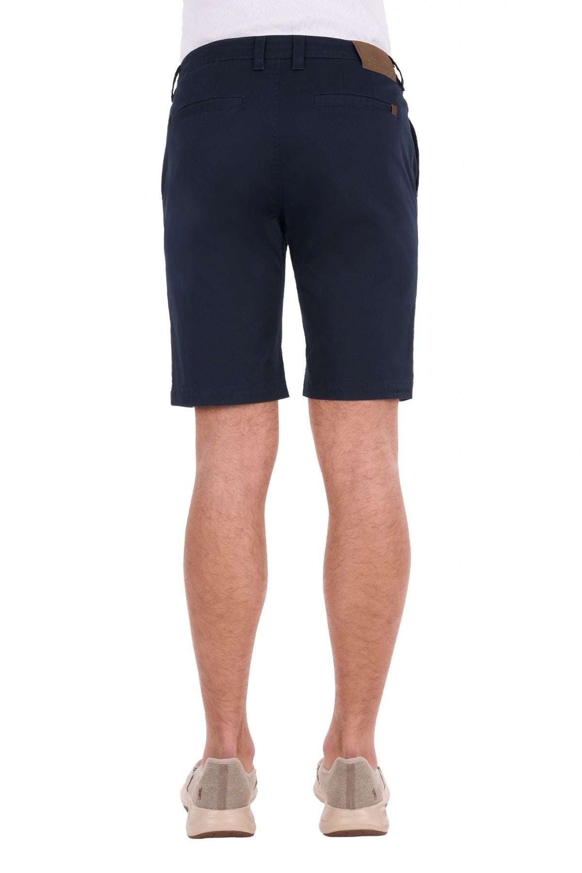 A person wearing navy blue Thomas Cook Harrison Shorts, made from stretch fabric with a vintage wash, paired with a white shirt and beige slip-on shoes, is standing facing away to display the back view of the shorts against a white background.