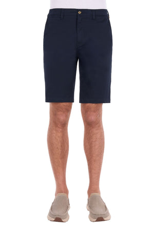 A man wearing the navy blue Thomas Cook Harrison Short, crafted from stretch fabric, paired with a white shirt and beige slip-on shoes, stands against a white background.