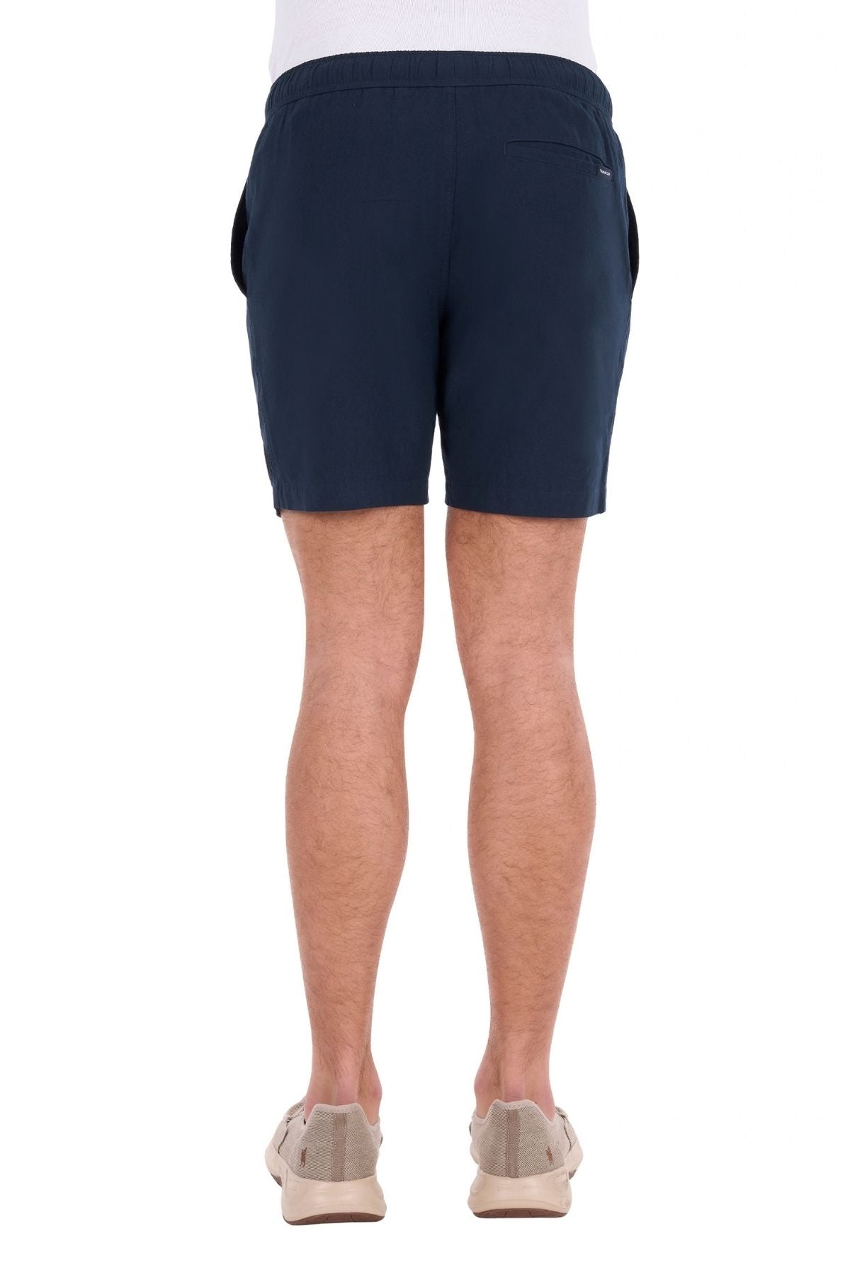 A person is standing facing away, wearing the Thomas Cook Jaxon Short in navy blue with an elastic waist, paired with beige slip-on shoes and a white top, against a plain white background.
