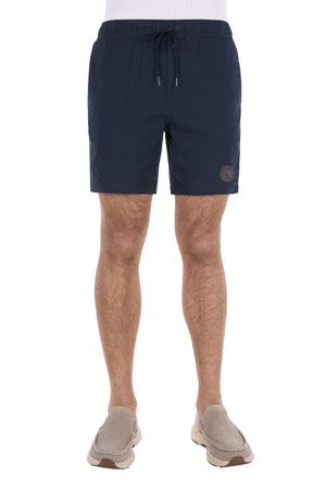 Wearing the Thomas Cook Jaxon Short in navy blue cotton linen with a drawstring waistband and paired with beige slip-on shoes, a person stands against a white background, effortlessly showcasing casual comfort.
