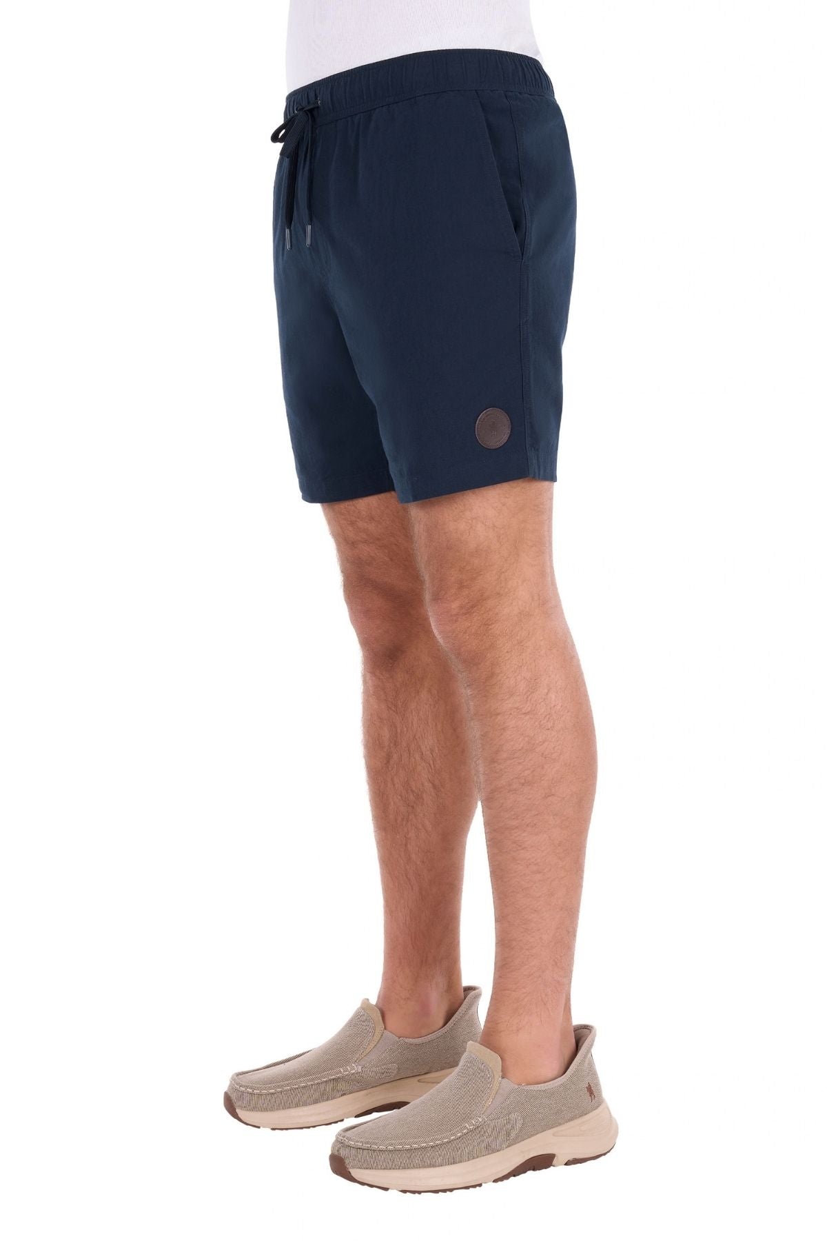 Wearing the Thomas Cook Jaxon Short in navy blue with an elastic waist for casual comfort, paired with a white shirt and beige slip-on shoes, the person stands against a plain white background.