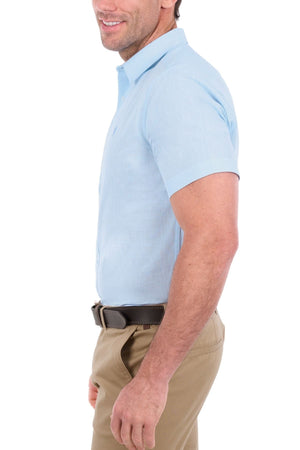 A man is standing sideways and smiling against a white background, wearing a light blue Thomas Cook Linen Short Sleeve Shirt, tan pants, and a dark belt.