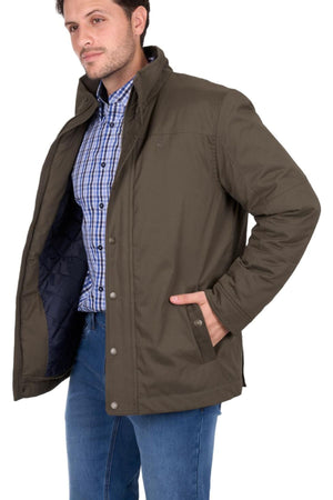 A man wears a brown Thomas Cook Matt Jacket midweight puffer over a blue and white plaid shirt and blue jeans, standing with one hand in his pocket and the other holding open his jacket, facing slightly to the side.