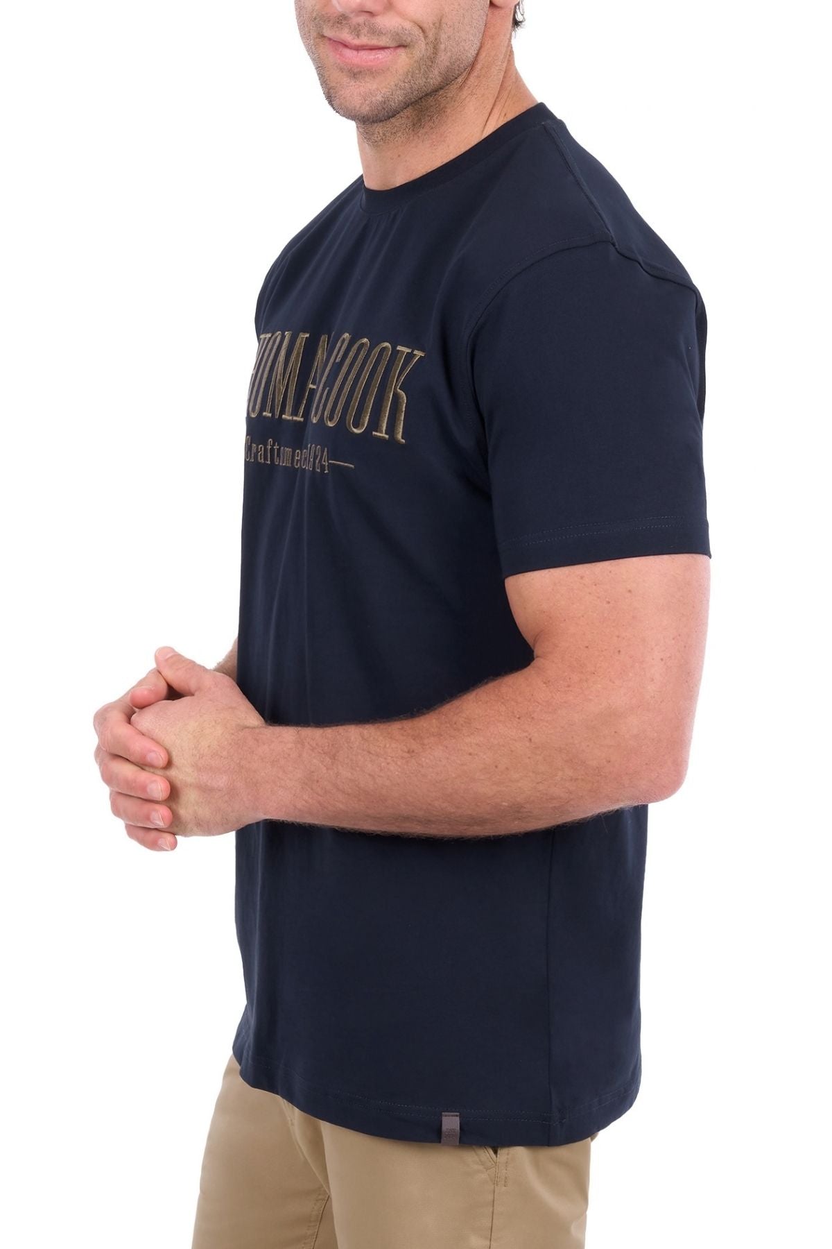 A man stands with his hands clasped, facing slightly to the side against a white background. He is wearing the Thomas Cook Nathan Tee in navy, which features gold chest embroidery, paired with tan pants.