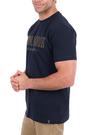 A man stands with his hands clasped, facing slightly to the side against a white background. He is wearing the Thomas Cook Nathan Tee in navy, which features gold chest embroidery, paired with tan pants.