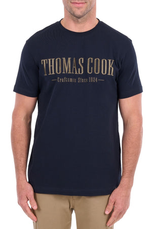 A man is shown from the waist up, facing forward with his arms relaxed at his sides, wearing a navy blue Thomas Cook Nathan Tee featuring “Thomas Cook Craftsmen Since 1924” embroidery on the chest and paired with beige pants.