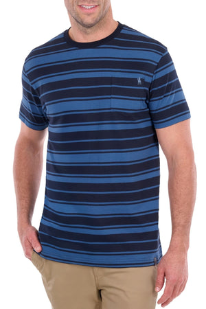A man wearing the Thomas Cook Pete Tee—a short-sleeve blue and navy striped shirt crafted from soft cotton jersey with a chest pocket—paired with tan pants, stands with one hand in his pocket and smiles.