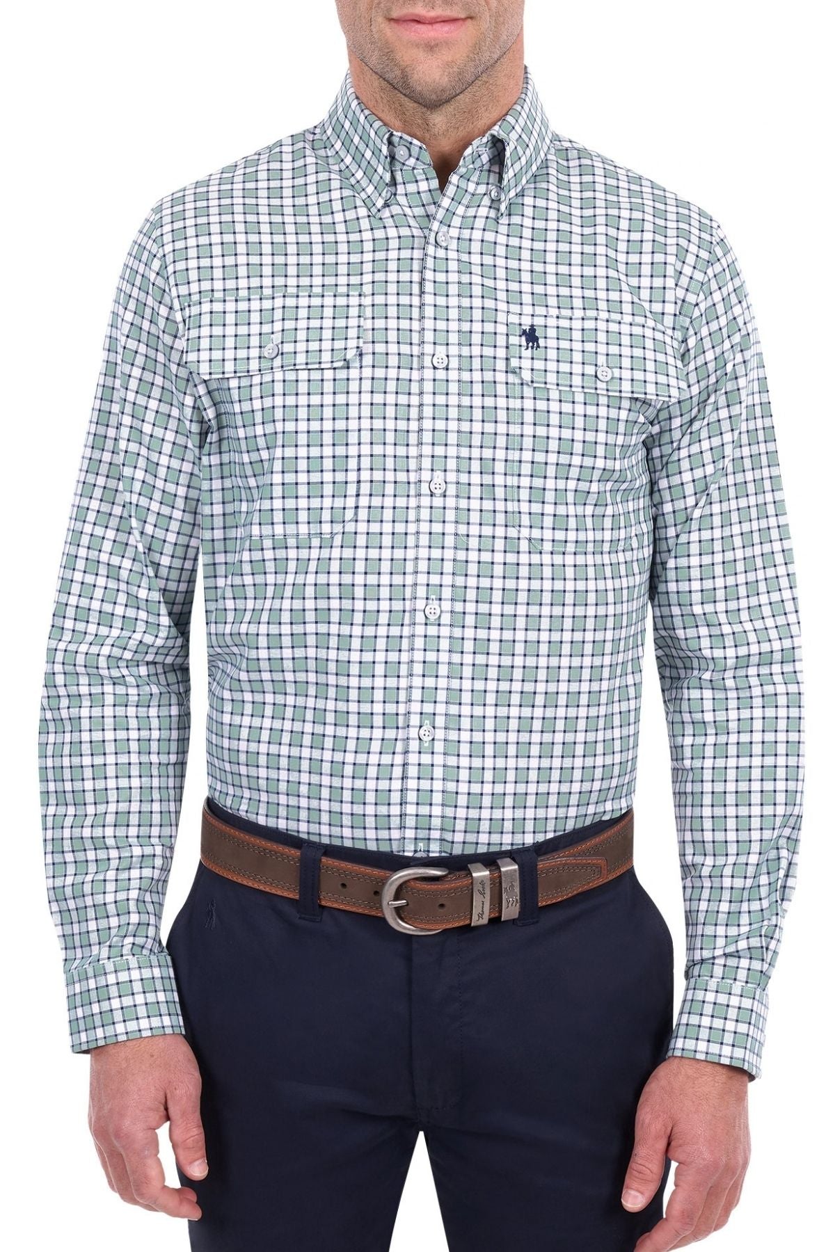 A man is wearing the Thomas Cook Rory Shirt, a green and white plaid cotton check shirt with long sleeves featuring front pockets and a small embroidered logo. He pairs it with dark pants and a brown belt, standing against a white background.