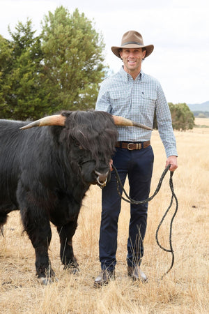 A man in a hat and the Thomas Cook Rory Shirt stands in a dry field, smiling and holding a rope attached to a large black cow with long horns and shaggy hair covering its eyes.