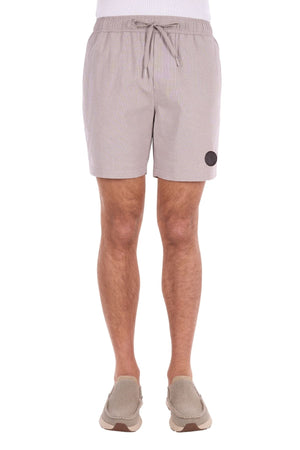 A person wearing light beige Thomas Cook Rowan Shorts, crafted from premium textured fabric with a drawstring and featuring a round black patch on the left leg, paired with matching beige slip-on shoes, stands against a white background.