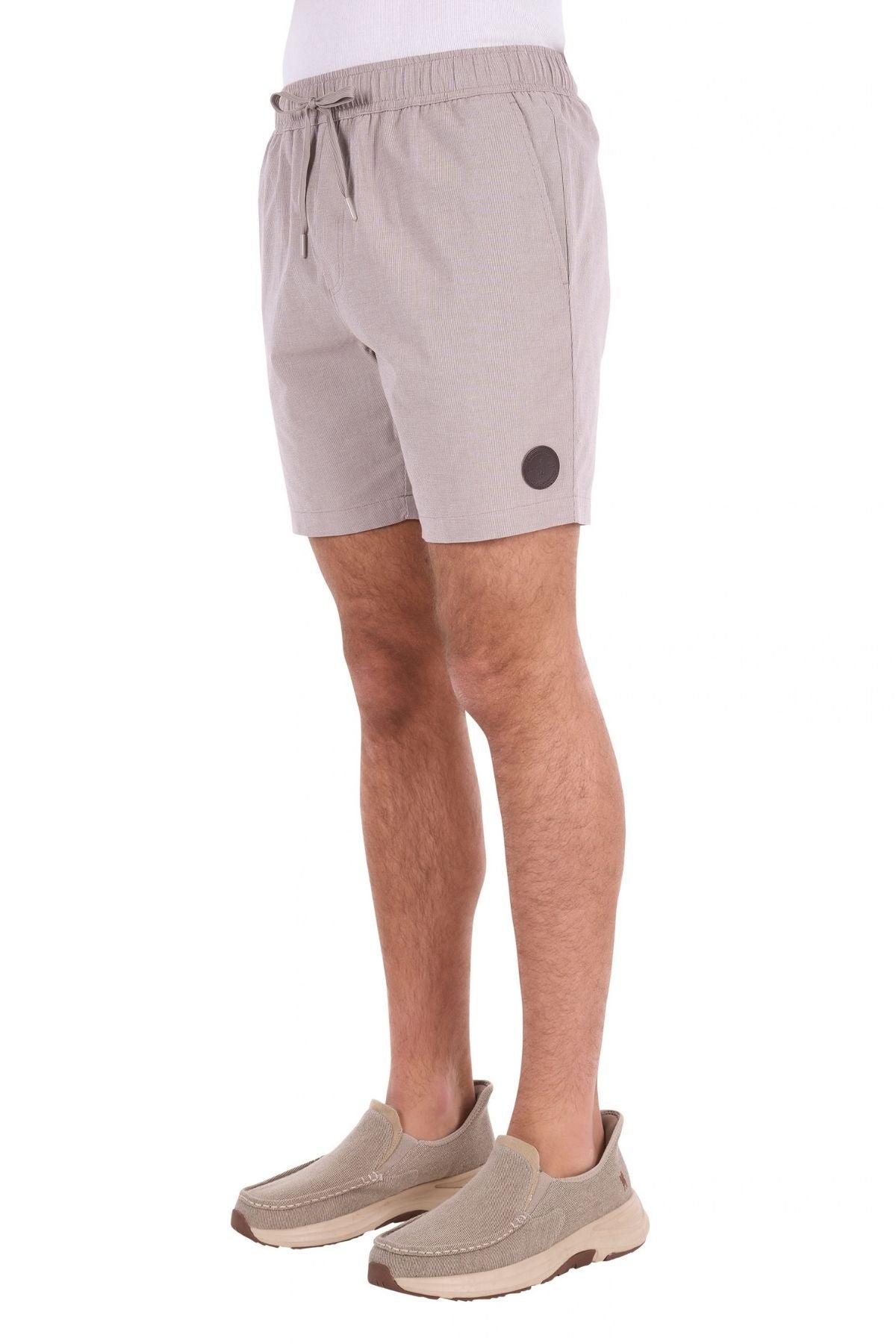 A man is wearing the Thomas Cook Rowan Short in light gray, made from premium textured fabric enhanced with stretch technology and featuring a drawstring waist and a black circular patch on the left leg. He pairs the shorts with beige slip-on shoes and stands against a white background.