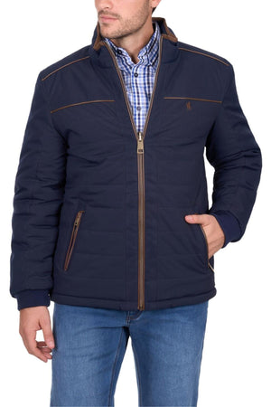 A man wears a navy blue Thomas Cook Toby Reversible Jacket with brown accents over a blue plaid shirt and blue jeans, standing with one hand in his pocket against a white background.