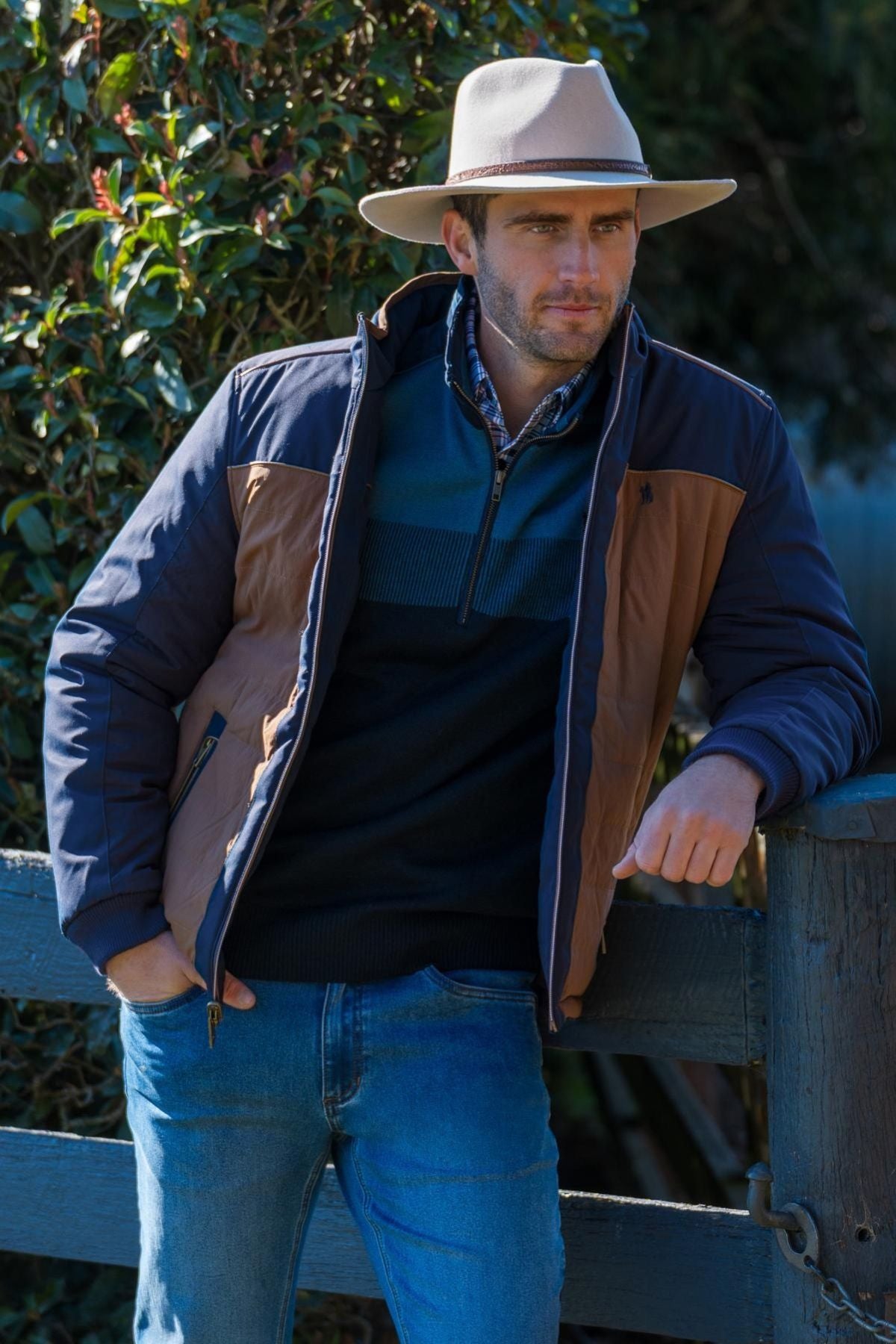 A man in a tan wide-brimmed hat, dark sweater, blue jeans, and the Thomas Cook Toby Reversible Jacket leans casually against a wooden fence with greenery behind him.