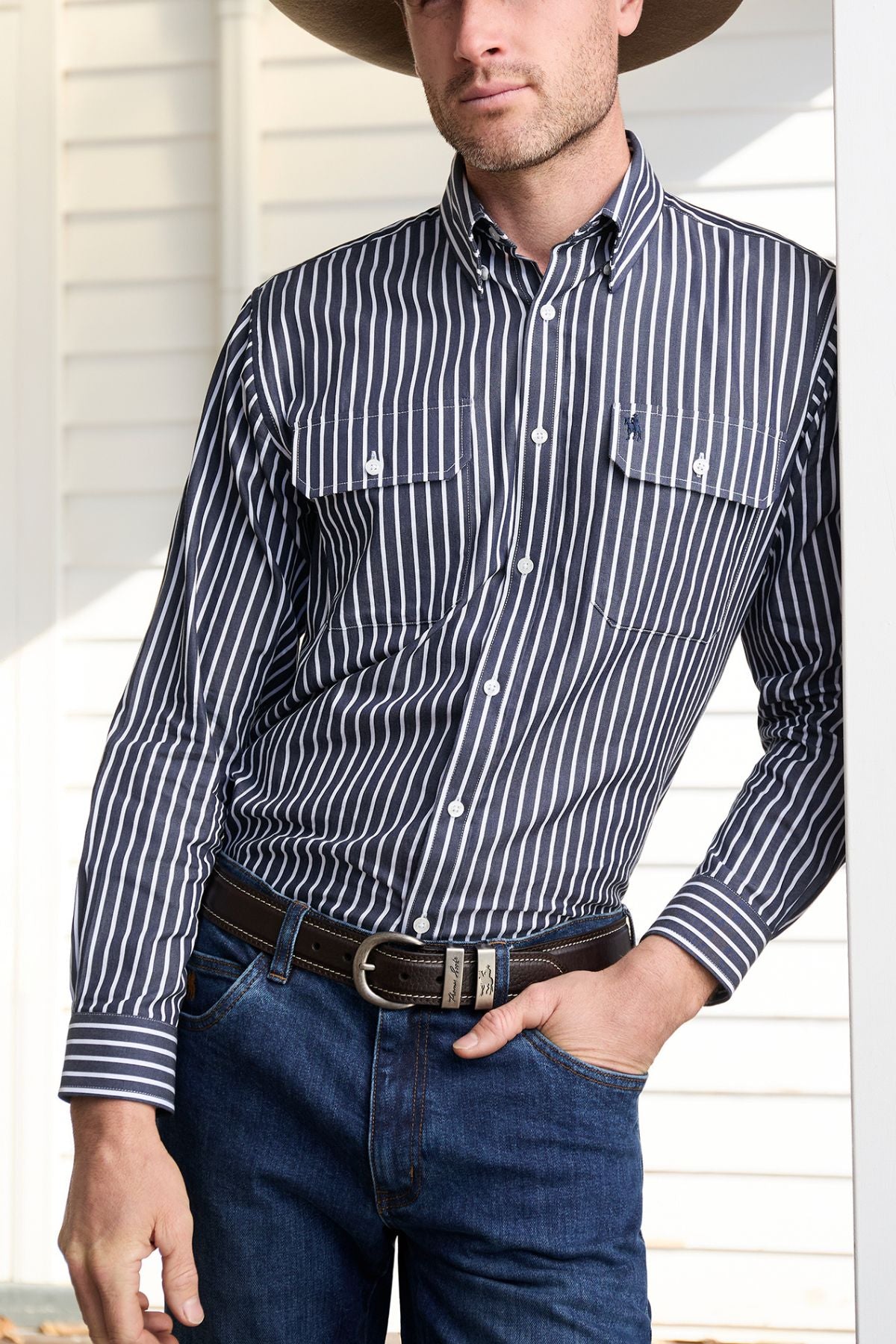 A man wearing a Thomas Cook Winton Shirt—a striped cotton shirt with double chest pockets—plus a wide-brimmed hat, dark belt, and blue jeans stands leaning against a white wall, his head partially out of frame.