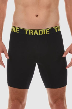 A person models black Tradie Big Fella Long Leg Trunks, featuring a yellow waistband with "TRADIE" in bold, made of stretch cotton for comfort and durability, set against a plain white background.