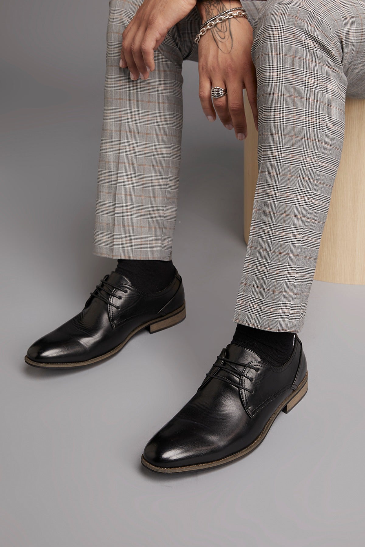 A person in grey plaid dress pants, black socks, and the UNcut Rhine Shoe (non-leather upper) sits on a light stool, hand on knee with rings and bracelets—a refined formal look.