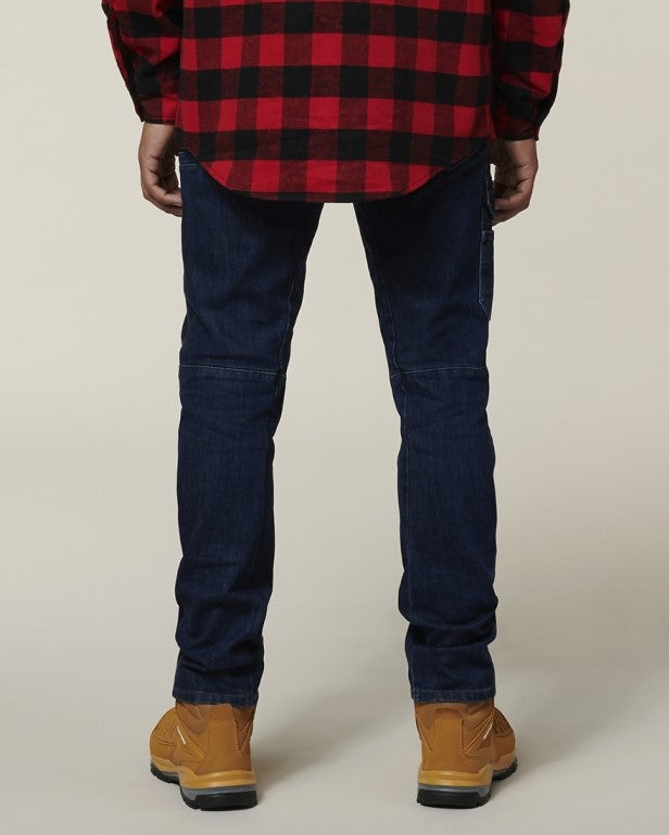 A person, facing away, wears a red and black checkered shirt, tan boots, and dark blue Hard Yakka Dyneema Tough Jeans against a plain light background.