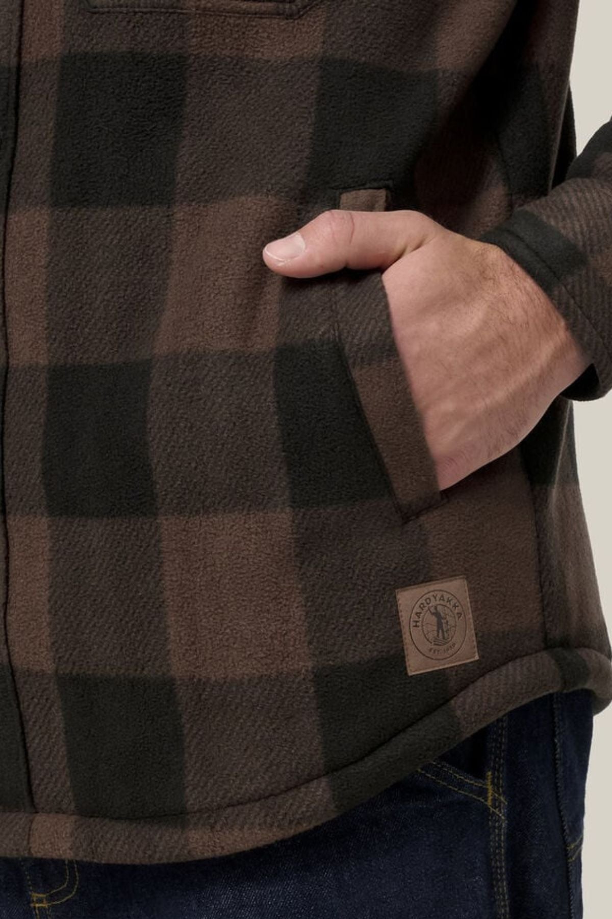 A person wears the Hard Yakka Legends Sherpa with FREE Beanie— a brown and black plaid flannel shirt with fleece lining, a small rectangular logo patch, and pairs it with dark blue jeans, right hand in the pocket.