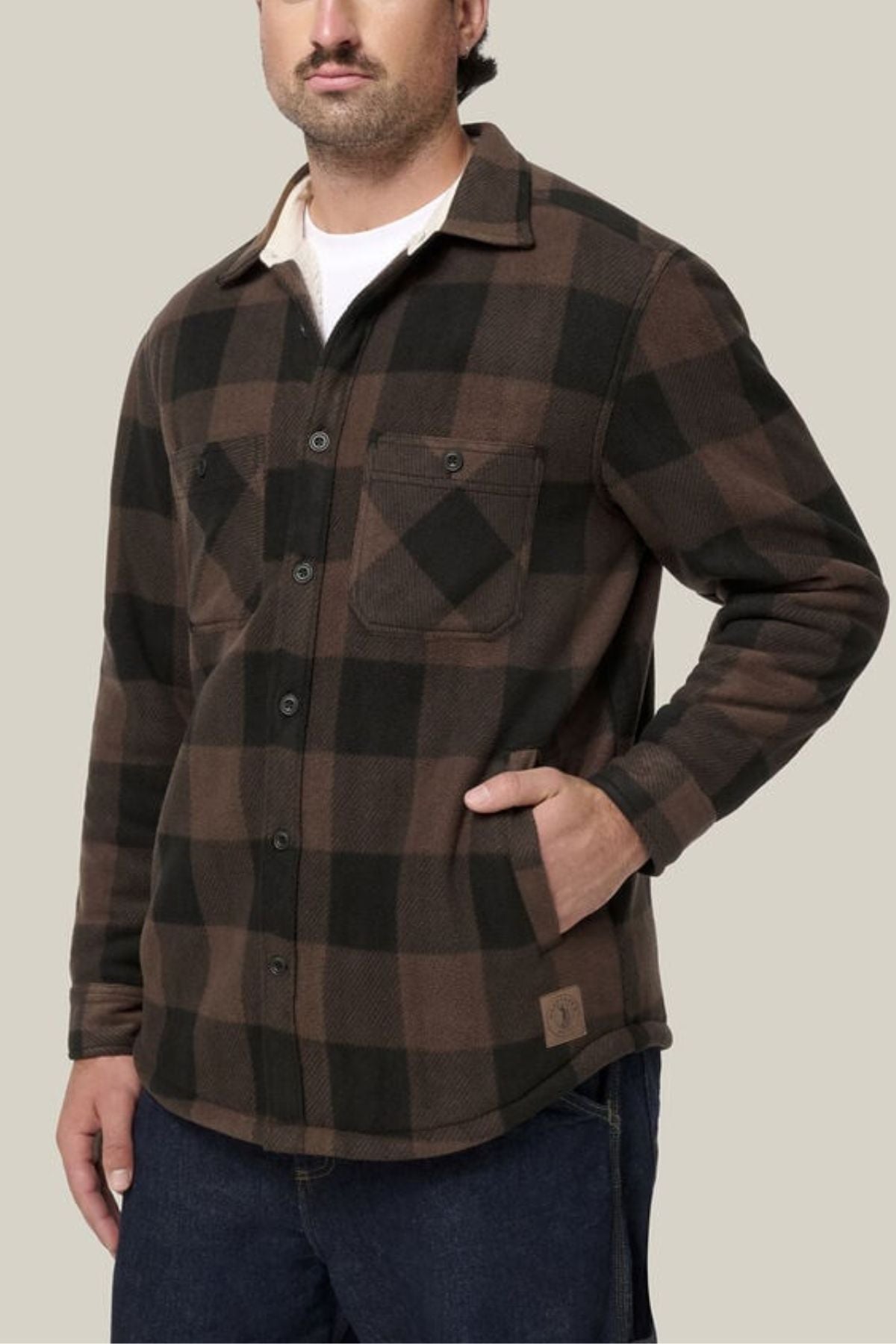 A man wearing a Hard Yakka Legends Sherpa with FREE Beanie, dark blue jeans, and a white undershirt stands with one hand in his pocket against a plain light background.