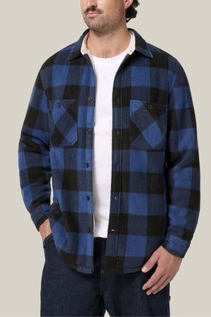 A man in a Hard Yakka Legends Sherpa with FREE Beanie, blue and black check flannel shirt, layered over a white t-shirt and dark jeans, stands with one hand in his pocket against a plain beige background.