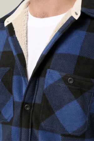 A close-up of a person wearing the Hard Yakka Legends Sherpa with FREE Beanie—blue and black plaid shirt with black buttons, cream fleece lining for thermal insulation, and layered over a white T-shirt with visible collar lining.
